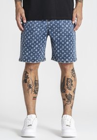 Mann trägt blaue, gemusterte Jeans-Shorts, weiße Sneaker und schwarzes Hemd; Tätowierungen einer Spinne, einer Frau und weiterer Motive an beiden Beinen sichtbar.