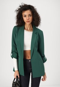 Žena s kovrčavom kosom, odjevena u zeleni sako, bijelu kratku majicu i tamne traperice, drži crnu torbicu na jednobojnoj pozadini.