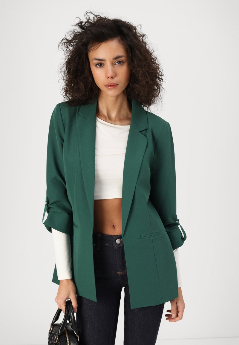 Blazer vert avec un design ajusté, des revers structurés et des manches retroussées. Associé à un crop top blanc et un jean foncé. Tissu texturé.
