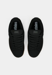 Μαύρα αθλητικά παπούτσια με μαλακό υφασμάτινο άνω μέρος, κορδόνια, στρογγυλή μύτη και καουτσούκ σόλα. Το λογότυπο είναι εμφανώς τοποθετημένο στη γλώσσα.