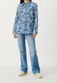 Person wearing a blue floral button-up shirt, light blue flare jeans, and white sneakers, standing against a plain light background.