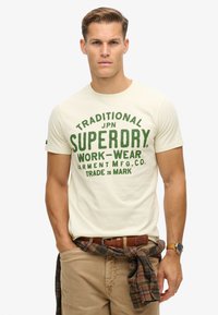 Camiseta beige con estampado verde que dice "Ropa de trabajo Superdry tradicional". Tiene mangas cortas y un cuello redondo. Se lleva con pantalones beige y una camisa de cuadros atada a la cintura.