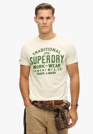 Beige T-shirt med grøn print med teksten "Traditional Superdry Workwear." Har korte ærmer og rund hals. Bæres med beige bukser og en ternet skjorte bundet om taljen.