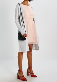 Světlounce šedý cardigan vrstvený přes světle růžové šaty s jemným plisé vzorem. Kombinováno s červenými lodičkami z lesklé kůže na blokovém podpatku.