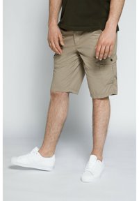 Pantaloni cargo beige con tasche laterali, realizzati in tessuto leggero, abbinati a sneaker bianche. I pantaloni hanno una vestibilità comoda e cuciture visibili.