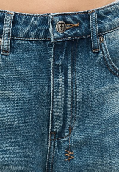 Κοντινό πλάνο στη μέση ενός μπλε τζιν παντελονιού με μεταλλικό κουμπί και εμφανή ραφή φερμουάρ.