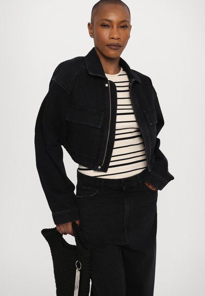 Chaqueta de mezclilla negra corta con dos bolsillos frontales, llevada sobre una camisa de rayas color crema. Se presentan jeans negros de cintura alta y un bolso negro texturizado.