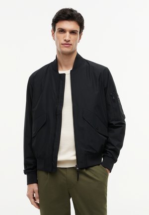 Homme portant une veste bomber noire sur un pull blanc et un pantalon vert olive, debout avec une main dans la poche sur fond blanc.
