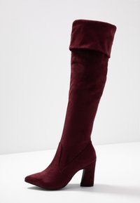 Botas burdeos hasta el muslo en suave ante con un puño vuelto, punta afilada y tacón grueso. Textura lisa sin herrajes visibles.