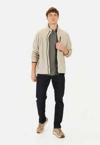 Beżowa polarowa kurtka z czarnym zamkiem, noszona na ciemnoszarym polo. Połączona z ciemnymi dżinsami i jasnobeżowymi trampkami.