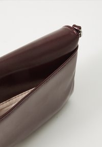 Pochette en cuir bordeaux avec une finition lisse, présentant une forme élancée et un accent subtil à l'ouverture. L'intérieur a une doublure claire.