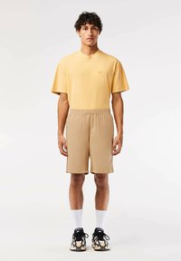 Camiseta de algodón amarilla de mangas cortas con un pequeño logo, combinada con pantalones cortos marrón claro. Llevada con zapatillas negras y blancas y calcetines blancos.