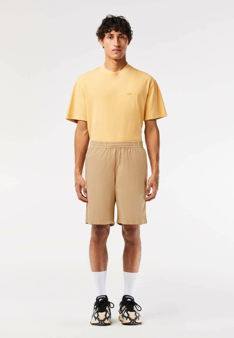 Camiseta de algodón amarilla de mangas cortas con un pequeño logo, combinada con pantalones cortos marrón claro. Llevada con zapatillas negras y blancas y calcetines blancos.