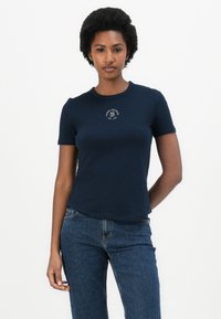 T-shirt bleu marine à manches courtes en coton avec un col rond, arborant un logo circulaire blanc sur la poitrine. Porté avec un jean bleu.