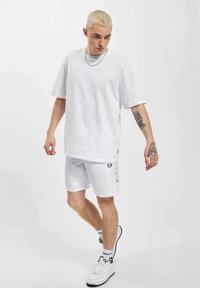 Hvit bomulls t-skjorte og shorts antrekk. T-skjorten har en løs passform. Shortsen har grafikk på sidene. Tilbehøret inkluderer en kjede og hvite joggesko.