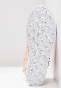 Semelle de chaussure en caoutchouc blanc embossée d'un motif répété du logo "GS" et du nom de la marque "GUESS", attachée à une chaussure beige à lacets.