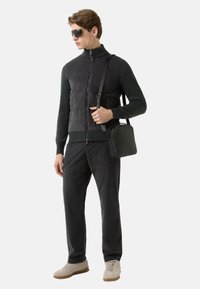 Sort vest med lynlås over en mørk trøje, kombineret med sorte bukser. En sort håndtaske bæres på tværs af kroppen. Beige sneakers fuldender looket.