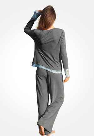 Mujer de pie descalza, vestida con una blusa de manga larga gris y pantalones con detalles de encaje azul claro en los puños y el dobladillo, vista desde atrás.