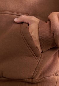 Hanorac maro cu buzunar tip kangaroo, manșete și tiv cu nervuri. Materialul pare moale, cu detalii de cusătură vizibile și ușor deteriorat.