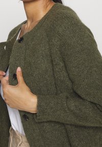 Olivgrön kofta med en texturerad stickad mönster, med två stora svarta knappar och ribbade muddarna, som erbjuder en avslappnad passform.