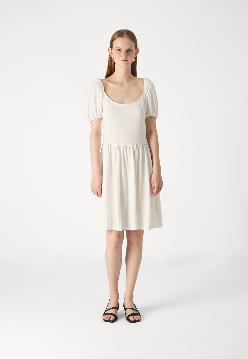 Robe en coton blanc avec des manches ballons courtes, un col rond, et une taille froncée, descendant en une jupe évasée. Portée avec des sandales noires.