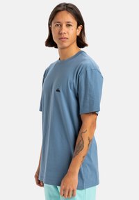 Camiseta de algodón azul claro con cuello redondo y mangas cortas, que presenta un pequeño logo negro en la parte izquierda del pecho. Textura suave.
