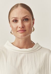 Blonde vrouw met groene ogen en sproeten draagt gouden ring oorbellen en een witte getextureerde top met een brede kraag, tegen een effen achtergrond.