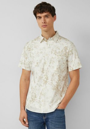 Mężczyzna w beżowo-białej, krótko rękawowej koszuli z subtelnym kwiatowym wzorem i niebieskich dżinsach, stojący na jednolitym jasnym tle.