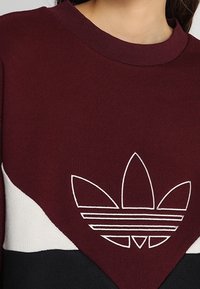 Detailní záběr na osobu, která má na sobě svetr v burgundské, bílé a černé barvě s velkým bílým logem Adidas trefoil na hrudi.