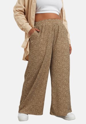 Kvinde iført løstsiddende, højtaljede brune bukser med hvide pletter, en hvid crop top, beige skjorte og hvide sneakers, hænderne i lommerne.