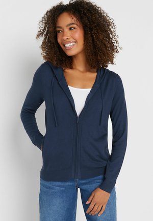 Lächelnde Frau mit lockigem Haar, die einen marineblauen Kapuzenpullover mit Reißverschluss über einem weißen Oberteil und blauen Jeans trägt, mit einer Hand in der Tasche stehend.