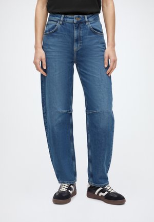 Person iført blå jeans med lige ben, sorte og hvide sneakers, stående mod en ensfarvet lys baggrund.