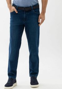 Jean en denim bleu foncé avec une coupe droite. Comprend des poches avant, une taille classique et une texture subtile. Porté avec une ceinture noire et des baskets.