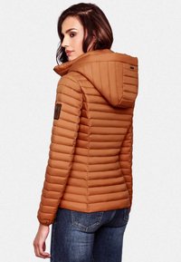 Ruda prigludusi pūkinė striukė su vertikaliu siuvinėtu raštu, pritvirtinta kapuce ir logotipo pleistru ant rankovės. Dėvima su tamsiai mėlynomis džinsomis.