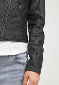 Zwarte leren motorjas met ritssluiting aan de voorkant, een textuurafwerking en nauwsluitende mouwen, gecombineerd met een wit shirt en lichtblauwe spijkerbroek.