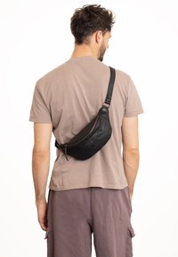 Zwarte leren schoudertas om crossbody te dragen, met een gebogen ontwerp, ritssluiting en een gladde textuur, gedragen door een persoon in een beige shirt.