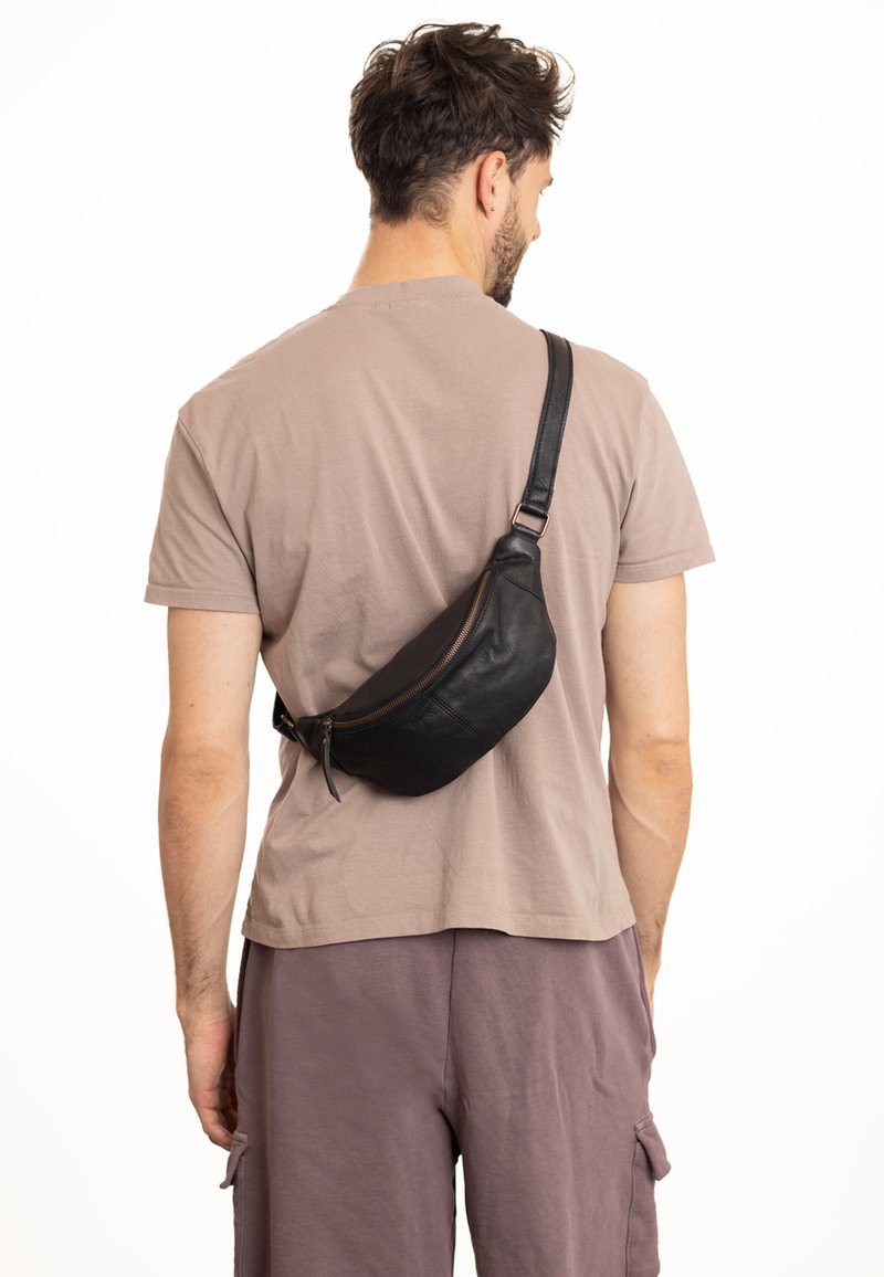 Zwarte leren schoudertas om crossbody te dragen, met een gebogen ontwerp, ritssluiting en een gladde textuur, gedragen door een persoon in een beige shirt.