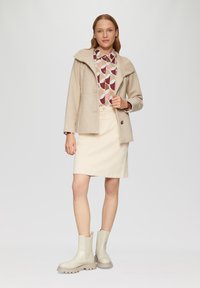 Beige wollen blend jasje met een opgerolde capuchon, gebloemde blouse met geometrische vormen in kastanjebruin, beige denim rok en lichtgrijze enkellaarsjes.