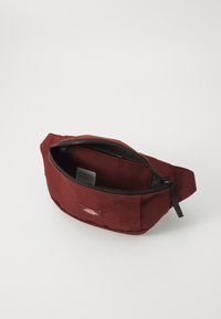 Burgundi színű szövet deréktáska fekete cipzárral, külső panelekkel és tágas belső térrel a tároláshoz.