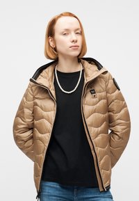 Νεαρό άτομο με ίσια κόκκινα μαλλιά φοράει μπεζ μπουφάν με καπιτονέ μοτίβο, μαύρο πουκάμισο, μαργαριταρένιο κολιέ και μπλε τζιν, στέκεται μπροστά σε λευκό φόντο.