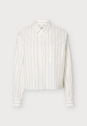 Femme debout portant une chemise blanche rayée avec des boutons et un pantalon large marron foncé avec des chaussures noires à bout carré.