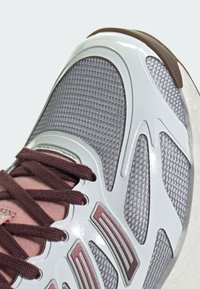 Scarpa sportiva con tomaia in mesh grigia, accenti in suede rosa e marrone, sovrapposizioni in plastica bianca e suola marrone scuro. I lacci sono marroni.