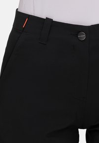 Close-up van een taille van een zwarte broek met een riemlus, een knoop met het label "Mammut" en een klein oranje stiksel aan de zijkant.