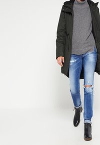 Tmavě zelená parka se stahovací šňůrkou v pase, přes šedý žebrovaný svetr, v kombinaci se světle modrými prodřenými džínami a černými kotníkovými botami.