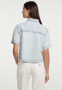 Helle blaue Denim-Jacke mit kurzen Ärmeln und einem cropped Design, sichtbaren Nähten und einem Kragen. Über weißen Hosen getragen, zeigt eine Rückansicht.