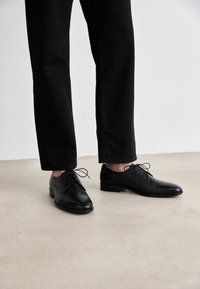 Black leather shoes with rounded toes and thin laces, worn with black pants. Minimalistic design with a low heel and smooth texture.