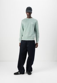Sudadera verde claro con manchas, pantalones anchos de mezclilla oscura, gorra negra y zapatillas negras. Look casual con detalle del logo en la sudadera.
