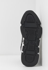 Semelle d'une basket noir et blanc avec un motif de bande de roulement géométrique et une indication de taille "37", positionnée verticalement contre un mur et un sol blancs.