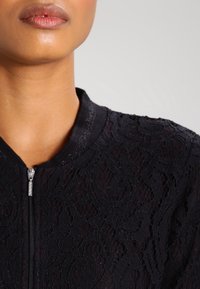 Bomber jacket in pizzo nero con motivo floreale strutturato, chiusura con zip e colletto a coste. Il tessuto appare leggero ed elegante.