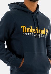 Marineblauwe katoenen hoodie met "Timberland Established 1973" in gele borduursel, buidelzak en ribboorden aan de mouwen en onderkant.